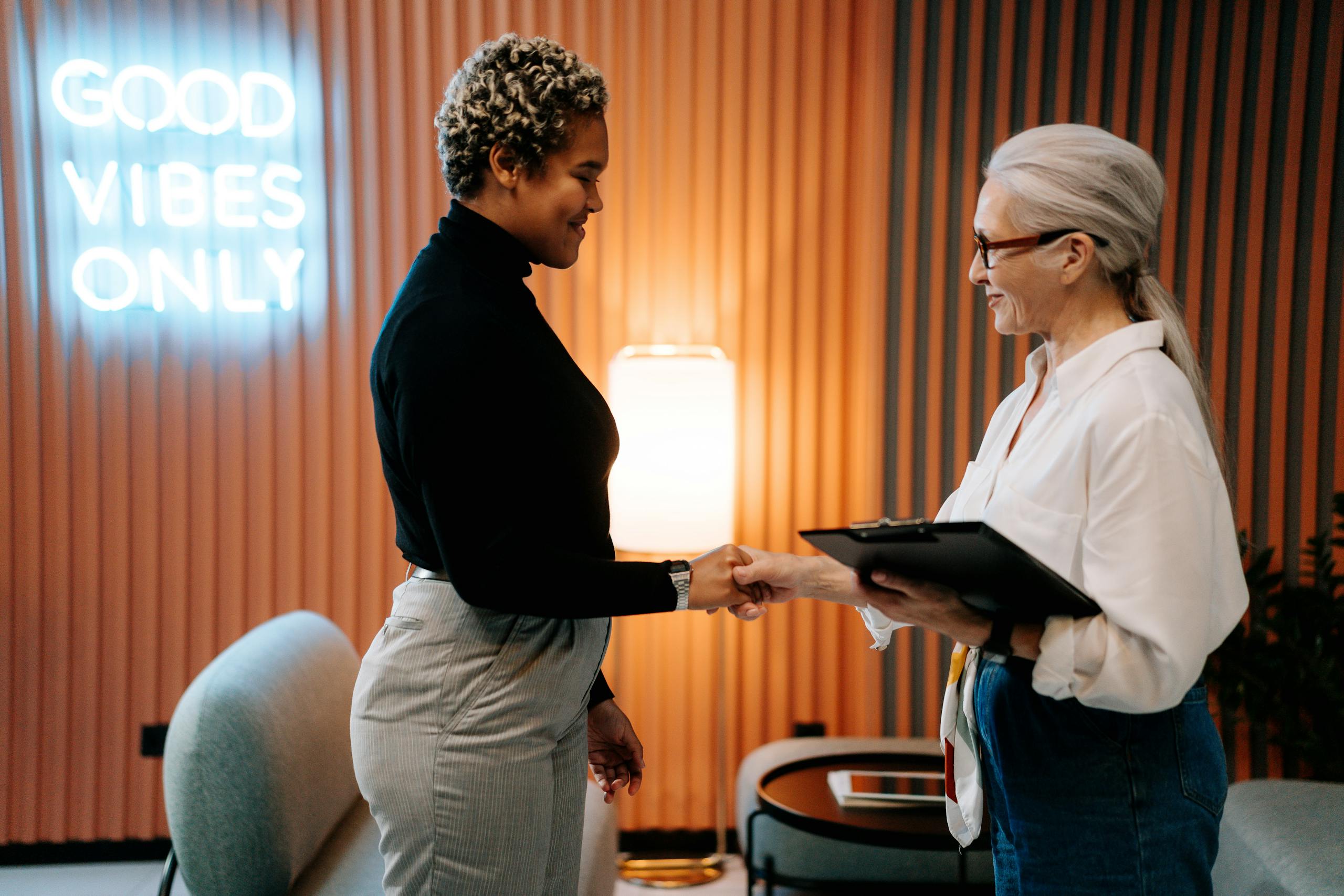 Two diverse businesswomen in a stylish office shaking hands under a 'Good Vibes Only' sign.